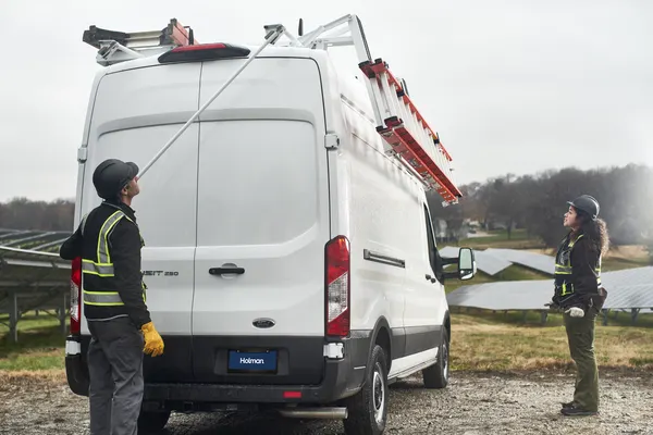 Two field technicians lowering an extension ladder from a drop-down ladder rack on a white Ford Transit
