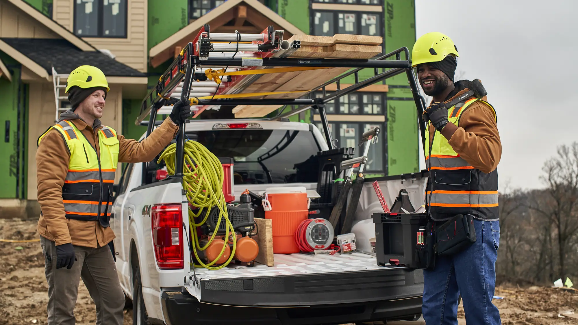 Two contractors unloading tools from a pickup fitted with a ladder rack at a home construction site