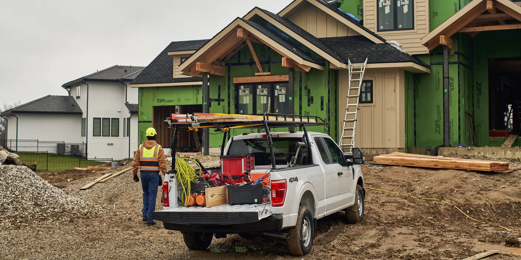 Contractor walking toward a white pickup loaded with tools and a ladder rack at a home construction site